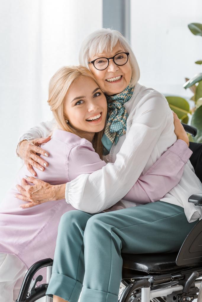 jovem mulher alegre abraçando mulher idosa em cadeira de rodas
  - Foto, Imagem