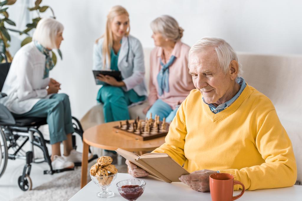 пожилой мужчина читает книгу, а женщина-врач осматривает пожилых женщин
 - Фото, изображение