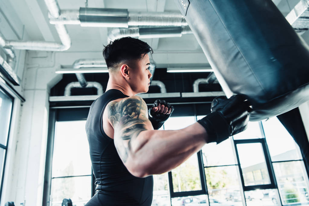 fiatal ázsiai sportember, boksz-kesztyűket visel és lyukasztó boksz zsák edzőteremben oldalnézetből - Fotó, kép