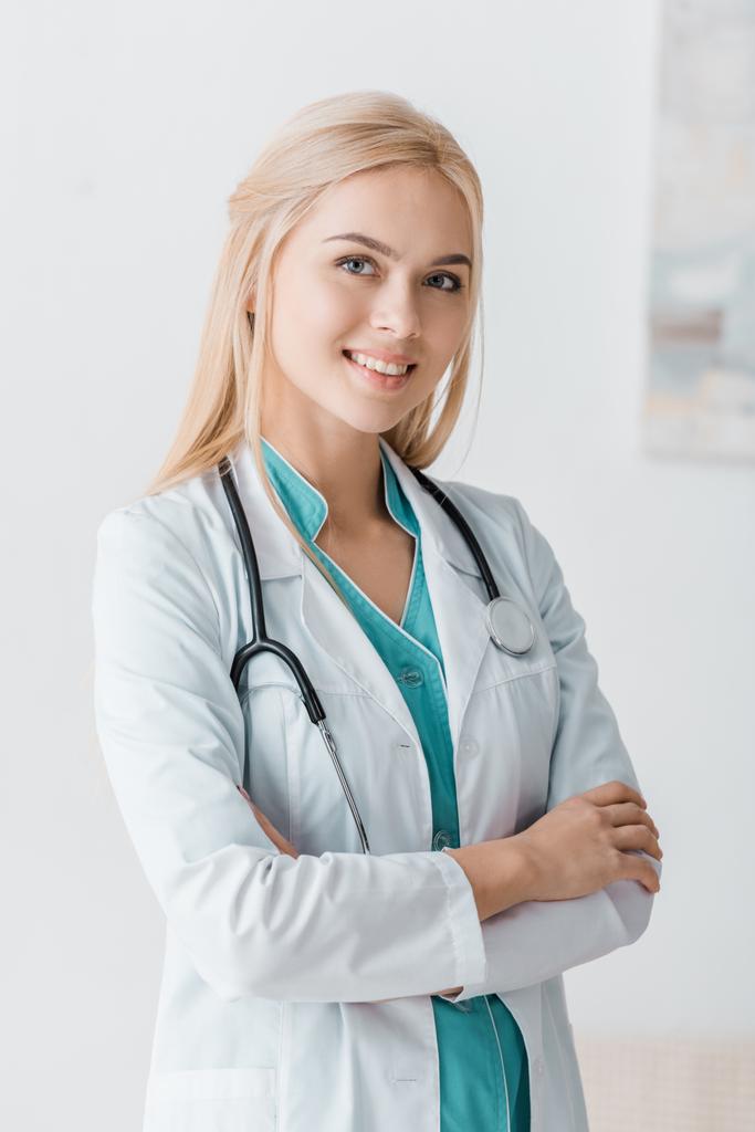χαμογελώντας νεαρή γυναίκα γιατρός με το στηθοσκόπιο στέκεται με τα χέρια σταυρωμένα στο ιατρείο  - Φωτογραφία, εικόνα