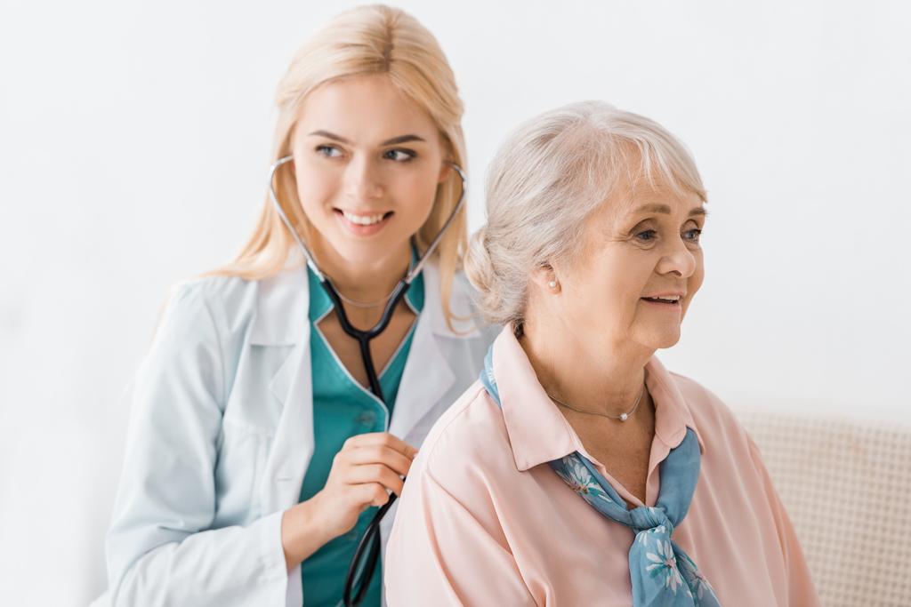 молодая улыбающаяся женщина-врач осматривает со стетоскопом пожилую женщину
 - Фото, изображение