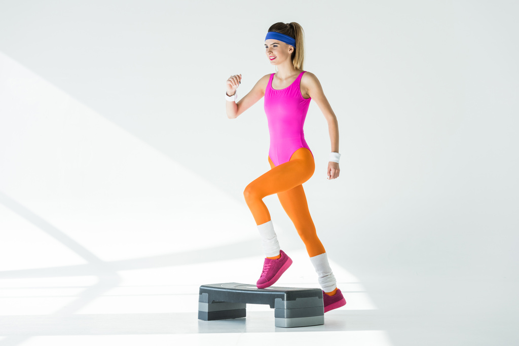 sorridente mulher desportiva exercitando-se na plataforma passo e olhando para longe em cinza
  - Foto, Imagem