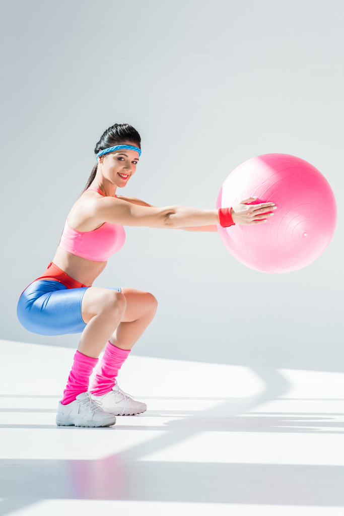 attraktive junge Frau trainiert mit Fitnessball und lächelt in die Kamera auf grau - Foto, Bild