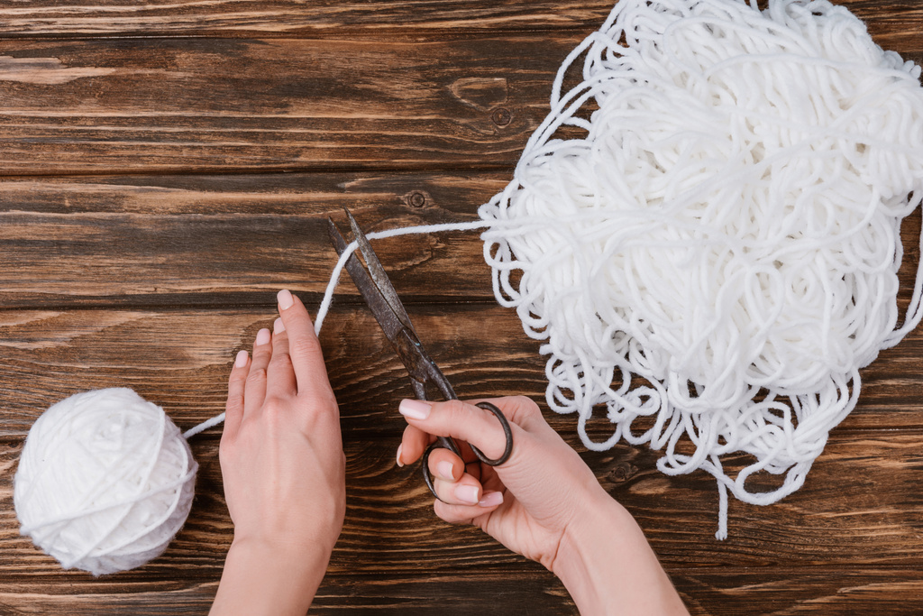 visão parcial da mulher cortando fio de tricô branco com tesoura na superfície de madeira
 - Foto, Imagem