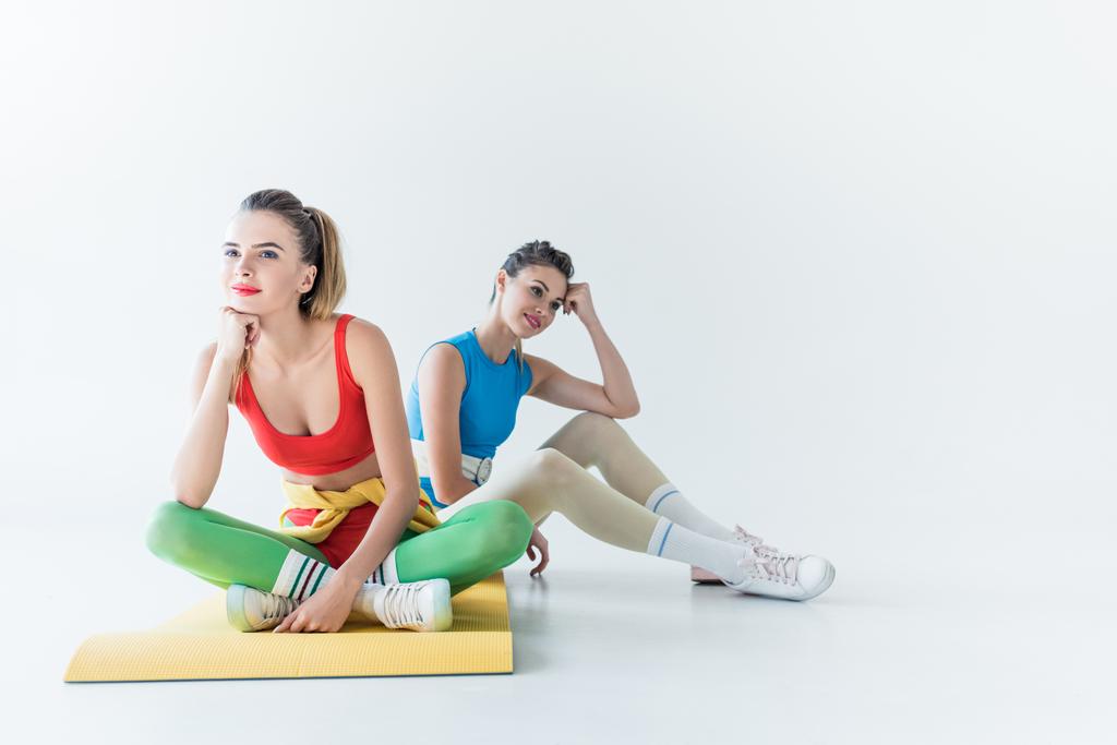 красивые улыбающиеся спортивные девушки, сидящие на коврике для йоги и отводящие взгляд от серого
 - Фото, изображение