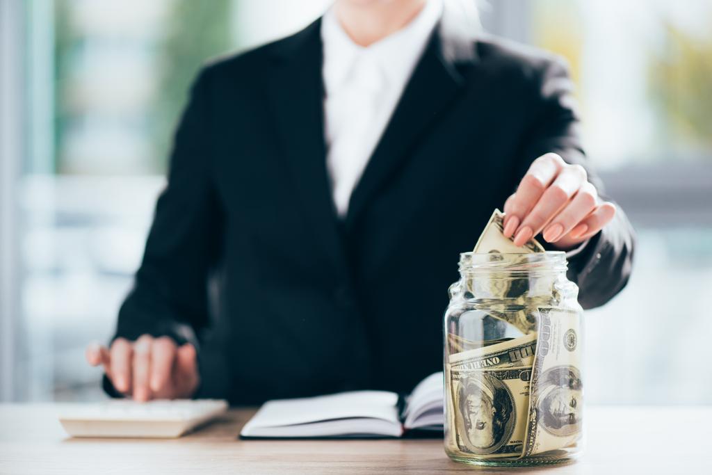Schnappschuss von Geschäftsfrau, die arbeitet und Dollarnoten in Glasgefäß steckt  - Foto, Bild