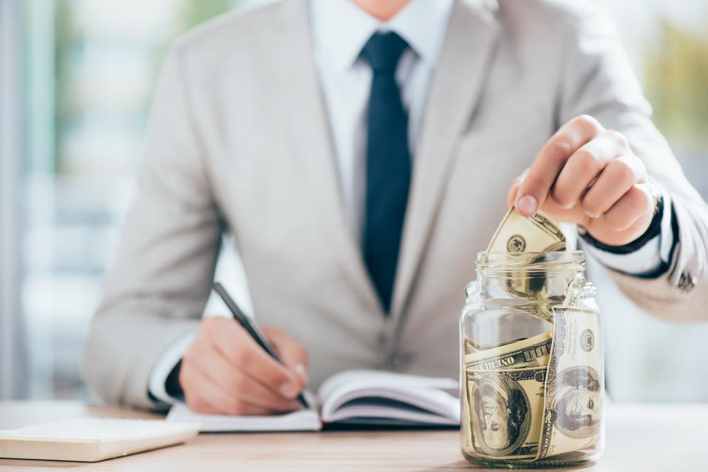 cropped shot of businessman working and putting dollar banknote in glass jar   - Fotografie, Obrázek