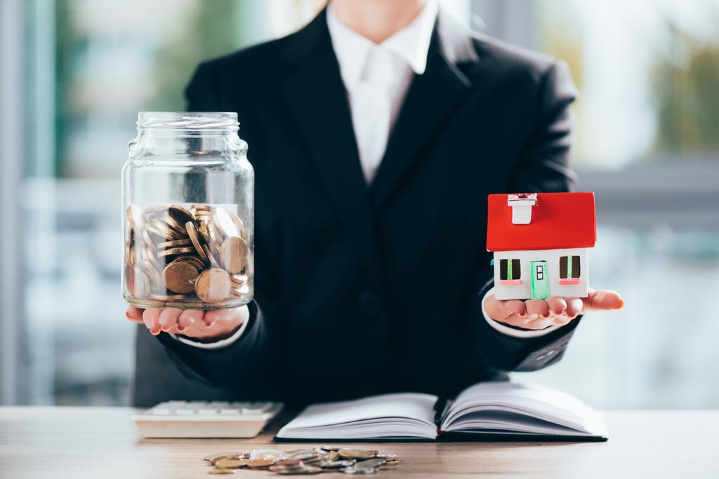 обрезанный снимок предпринимательницы, держащей стеклянную банку с монетами и моделью маленького дома
 - Фото, изображение