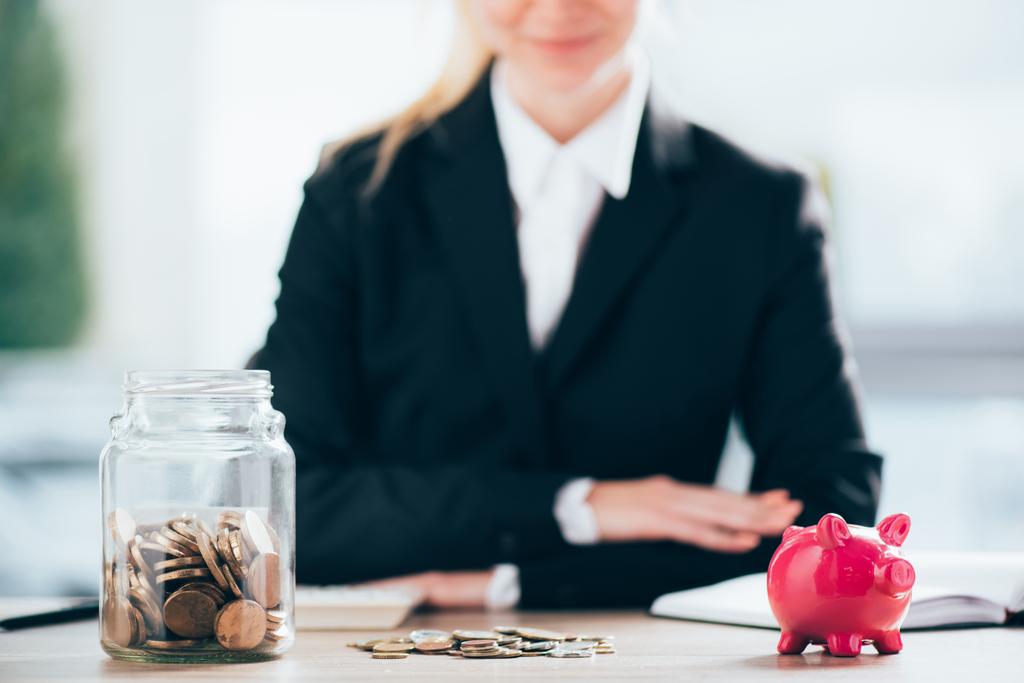 Nahaufnahme von rosa Sparschwein und Glasgefäß mit Münzen, lächelnde Geschäftsfrau dahinter - Foto, Bild