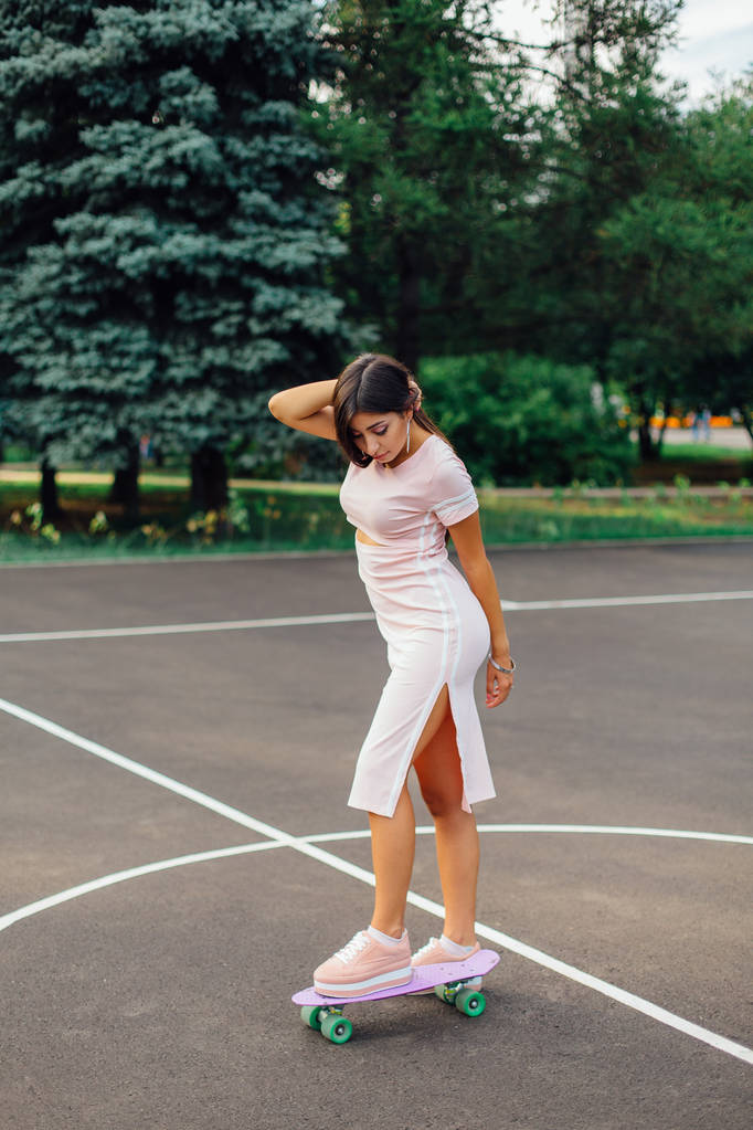 Portrait d'une charmante brune souriante debout sur son skateboard sur un terrain de basket-ball. Femme heureuse avec look tendance prenant une pause pendant le coucher du soleil
. - Photo, image