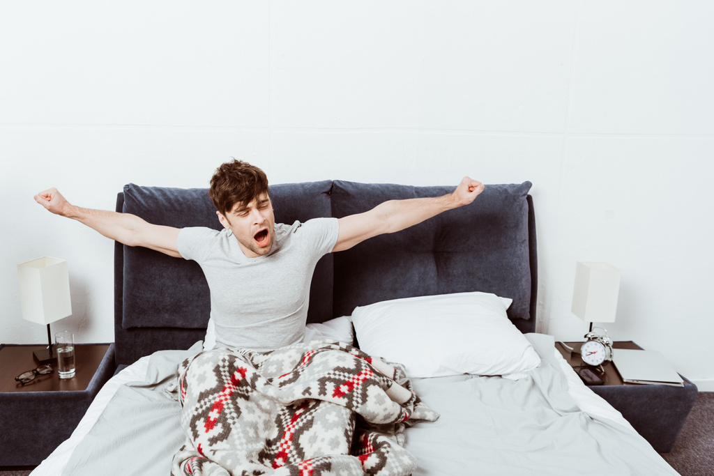 όμορφος νεαρός άνδρας χασμουρητό και εκτείνεται στο κρεβάτι κατά τη διάρκεια του χρόνου το πρωί στο σπίτι - Φωτογραφία, εικόνα