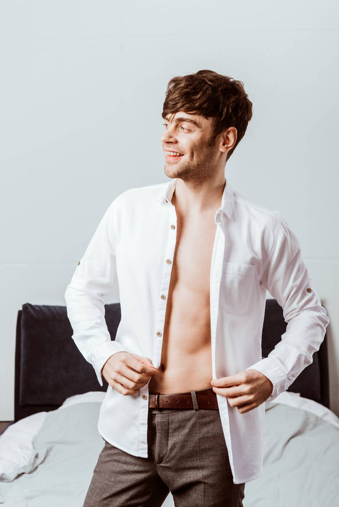 χαμογελαστός νεαρός επιχειρηματίας κουμπιά από λευκό πουκάμισο στο υπνοδωμάτιο στο σπίτι - Φωτογραφία, εικόνα