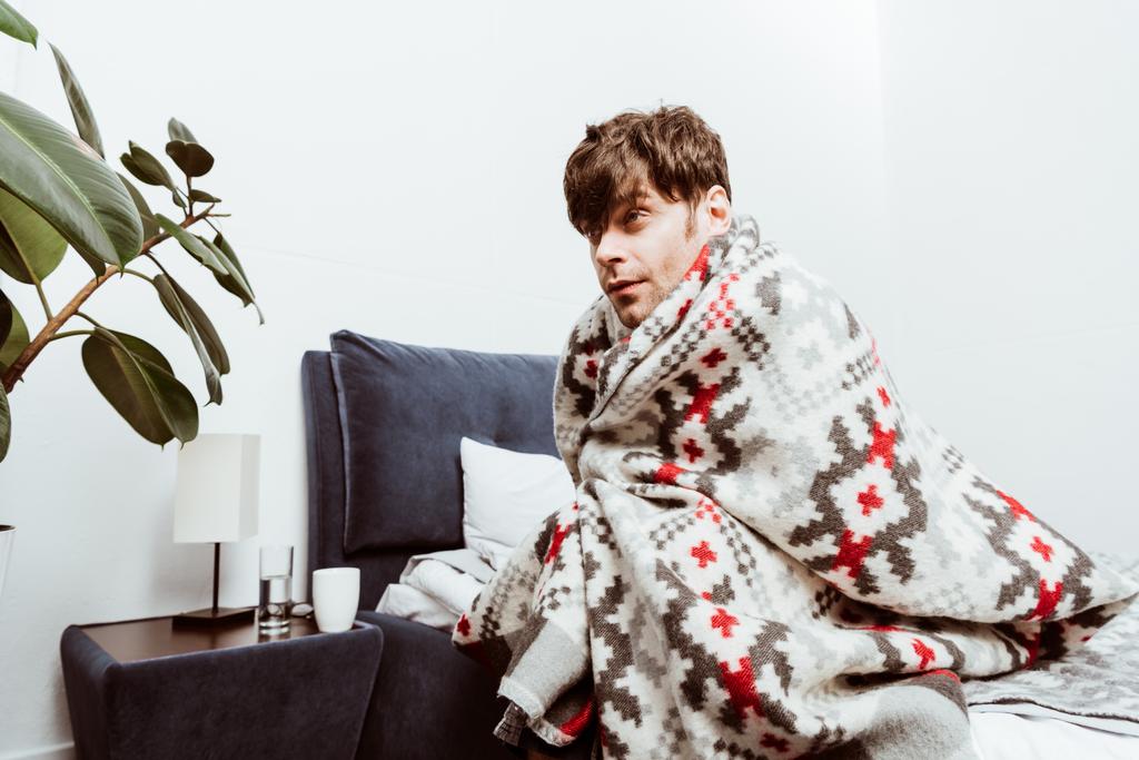 επιλεκτική εστίαση του άρρωστος νεαρός άνδρας τυλιγμένο σε κουβέρτα κάθεται στο κρεβάτι στο σπίτι - Φωτογραφία, εικόνα