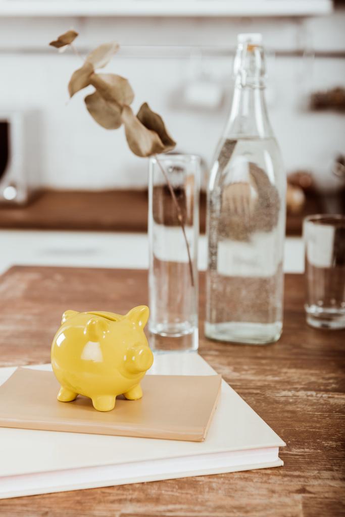Nahaufnahme des gelben Sparschweins auf Lehrbuch am Holztisch mit Wasserflasche und Vase mit Zweig  - Foto, Bild