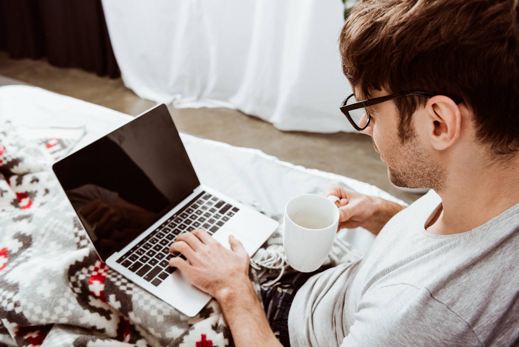 visão lateral do freelancer masculino com xícara de café trabalhando no laptop com tela em branco na cama em casa
 - Foto, Imagem