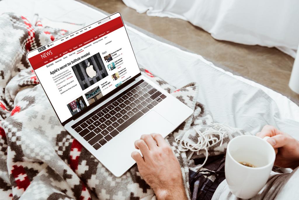 körülvágott kép az ember gazdaság csésze kávé és használ a laptop a bbc hír képernyőn az ágyban otthon - Fotó, kép