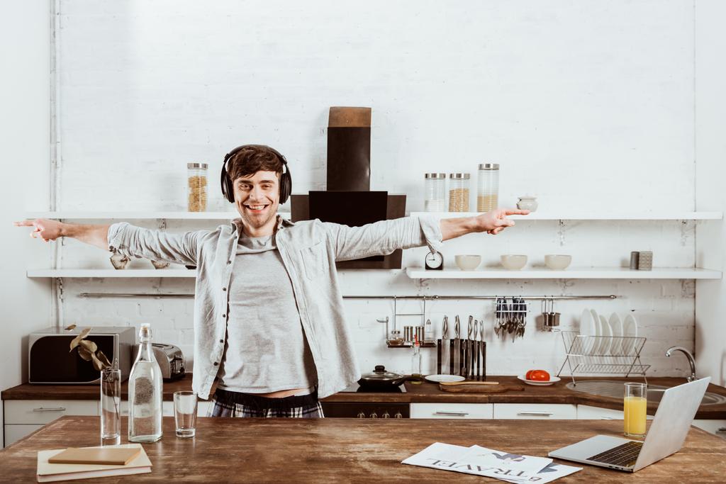 веселый мужчина фрилансер в наушниках, стоящий с широкими руками возле стола с ноутбуком на кухне дома
 - Фото, изображение