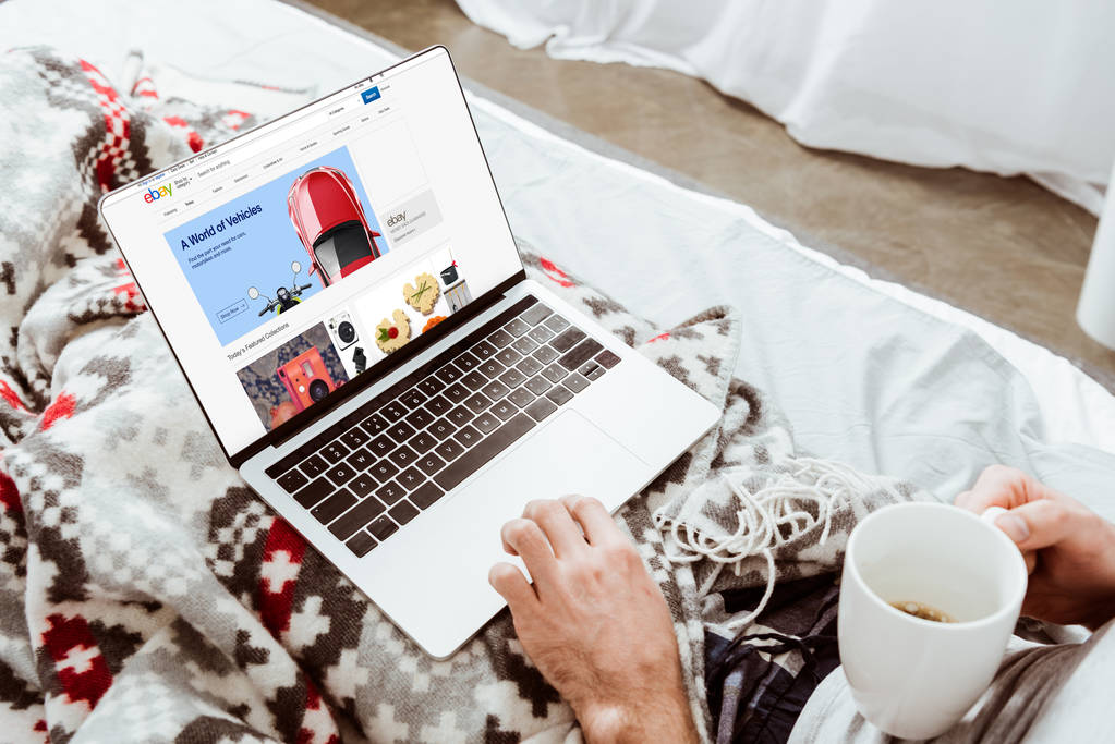 körülvágott kép az ember gazdaság csésze kávé és használ laptop-val ebay képernyőn az ágyban otthon - Fotó, kép