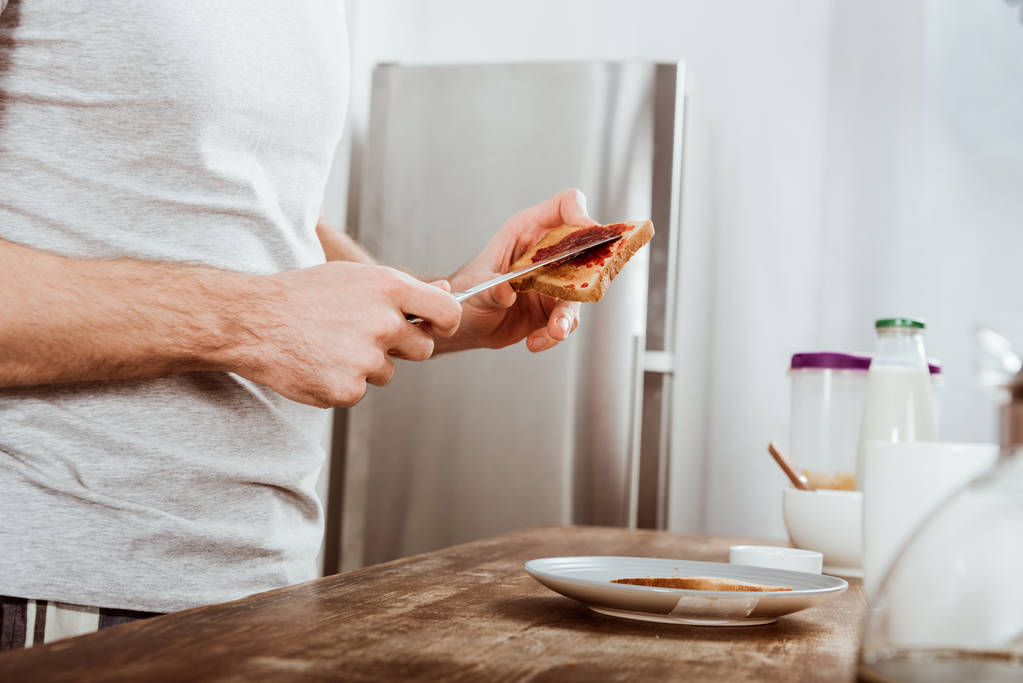 Teilansicht eines Mannes, der zu Hause in der Küche Toast nach Marmelade verteilt - Foto, Bild