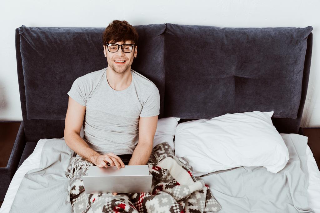 alegre freelancer masculino trabajando en el ordenador portátil y mirando a la cámara en la cama en casa
 - Foto, Imagen