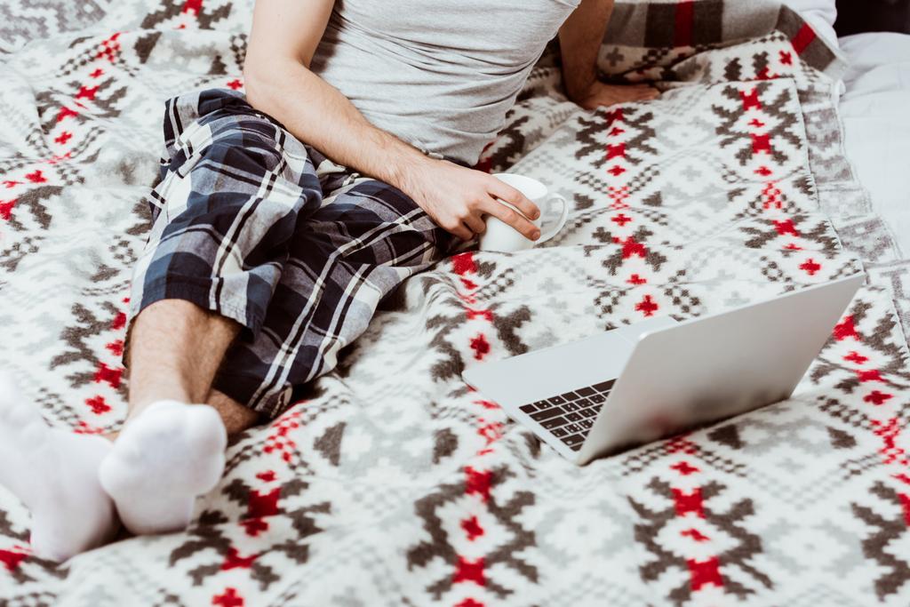 partial view of male freelancer with coffee cup sitting on bed with laptop at home - Photo, Image