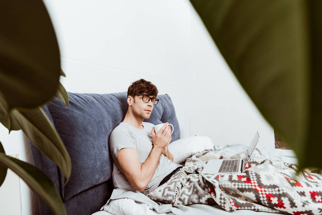 избирательный фокус задумчивый мужчина фрилансер с чашкой кофе сидя на кровати с ноутбуком дома
 - Фото, изображение