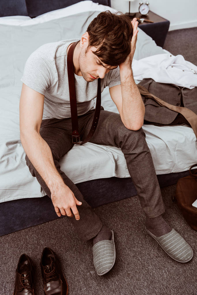 joven hombre de negocios con cinturón sobre el cuello sentado en la cama y con dolor de cabeza en casa
 - Foto, Imagen