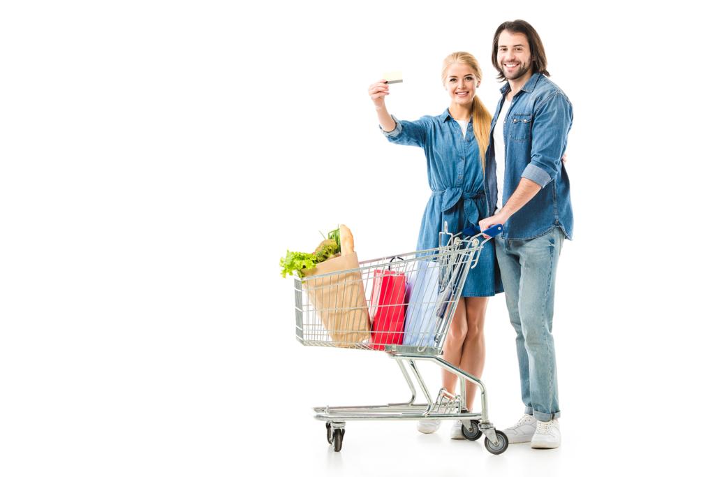 Preciosa pareja con carrito de compras lleno de bolsas que muestran la tarjeta de crédito aislado en blanco
 - Foto, Imagen
