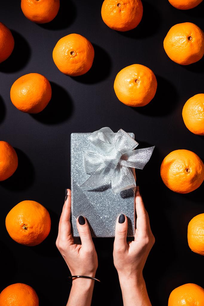vista parcial da mulher segurando caixa de presente de prata na mesa preta com tangerinas frescas ao redor
 - Foto, Imagem