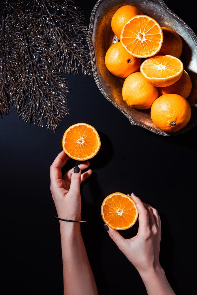 visão parcial da mulher segurando metades de tangerina na superfície preta com galho de prata decorativo e tangerinas em tigela de metal
 - Foto, Imagem