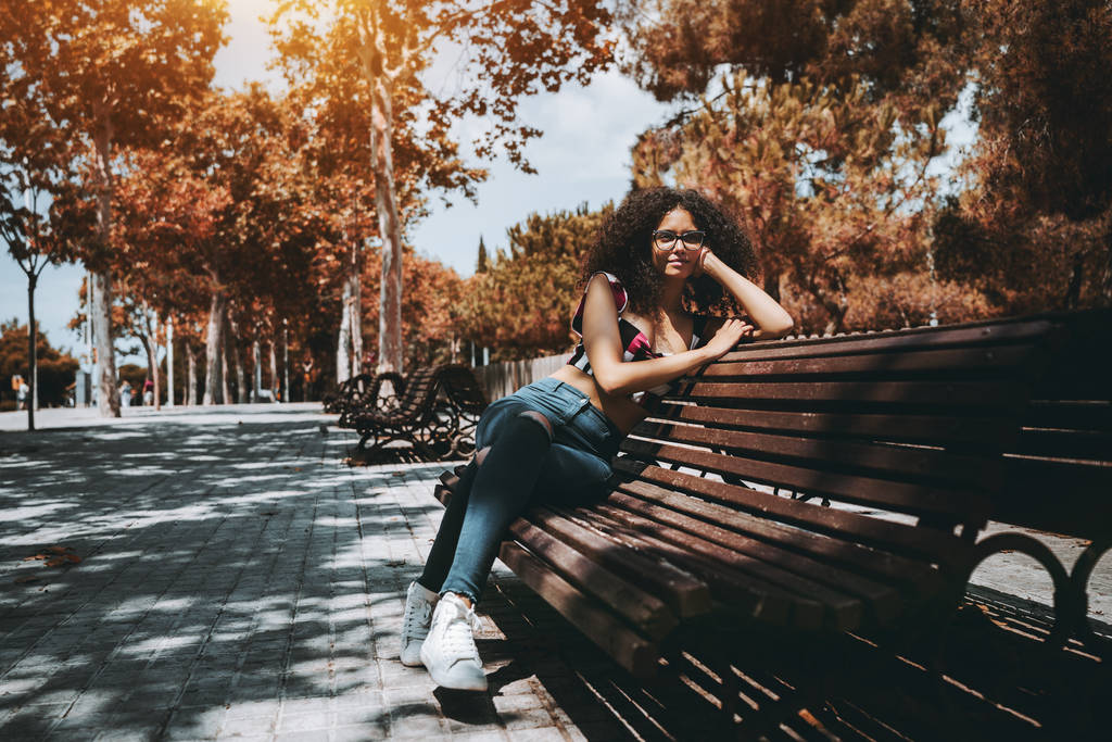 Czarujący młody kaukaski kobieta w okularach i z wielkogabarytowych kręcone włosy tęsknie patrzy gdzieś na zewnątrz siedząc na ławce na chodniku, a słoneczny; obszar przestrzeni kopii po lewej stronie - Zdjęcie, obraz