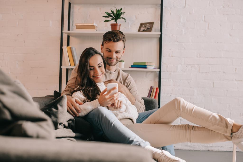 Jeune couple souriant utilisant un smartphone ensemble sur le canapé à la maison - Photo, image