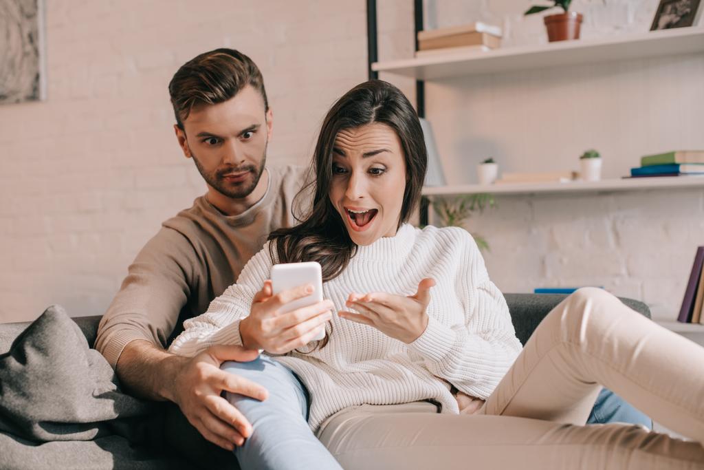 expressive young couple using smartphone together on couch at home - Photo, Image