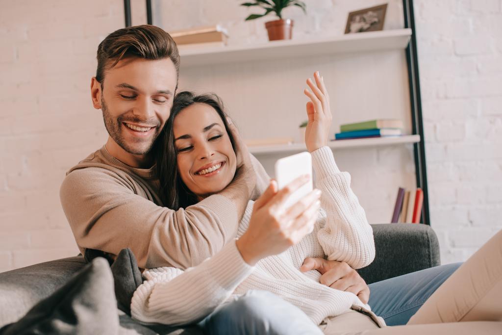Hymyilevä nuori pari käyttää älypuhelinta yhdessä sohvalla kotona - Valokuva, kuva