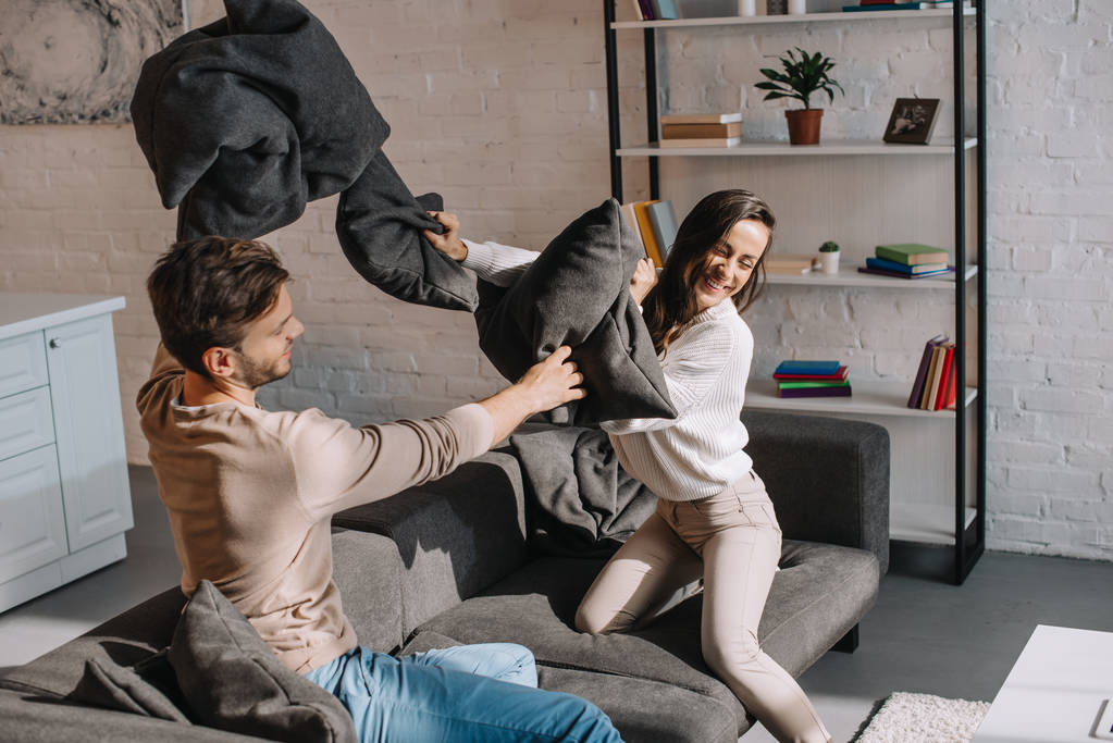 laughing young couple fighting with pillows on couch at home - Photo, Image