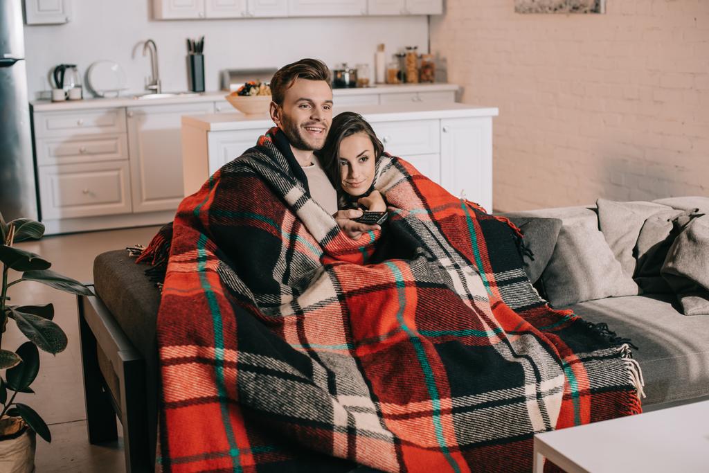 glückliches junges Paar, das es sich auf der Couch gemütlich macht und gemeinsam fernsieht, während es sich mit Plaid bedeckt - Foto, Bild
