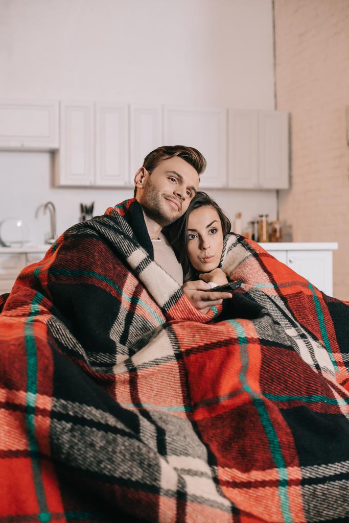 感情的な若いカップルがソファでリラックスと格子縞でカバーしながら一緒にテレビを見て - 写真・画像