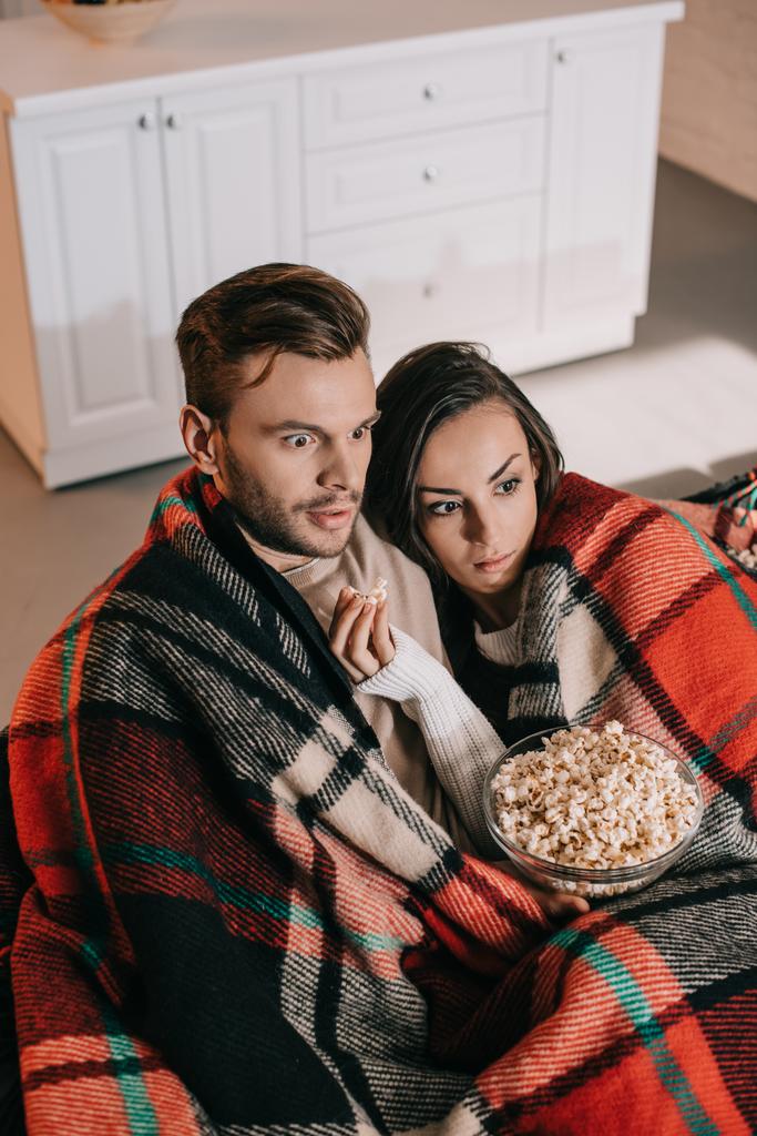 Blick aus der Vogelperspektive auf ein schockiertes junges Paar, das auf der Couch einen Film mit Popcorn anschaut und sich mit Plaid bedeckt - Foto, Bild