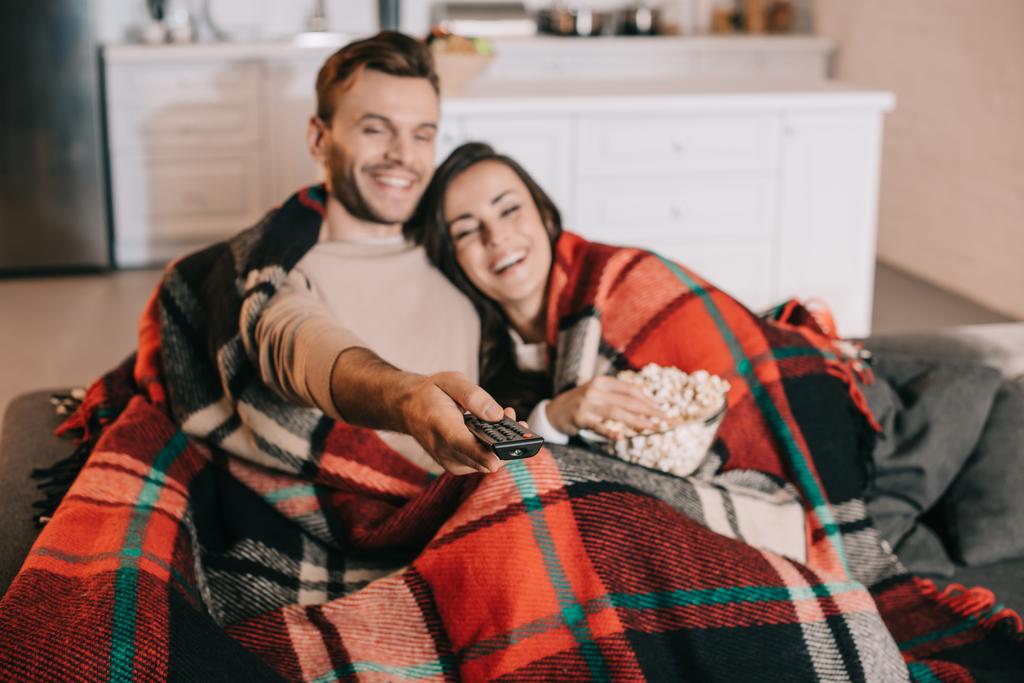 счастливая молодая пара смотрит телевизор с попкорном на диване и покрывает пледом
 - Фото, изображение