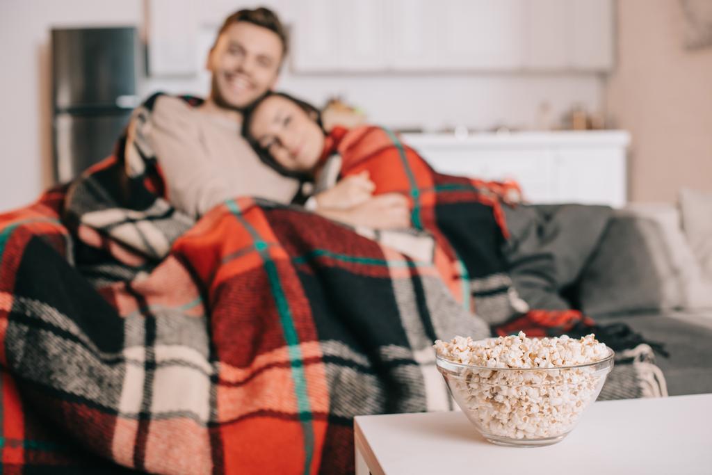 krásná mladá dvojice odpočinku na gauči a pokrývající s plaid s mísa popcornu na popředí - Fotografie, Obrázek