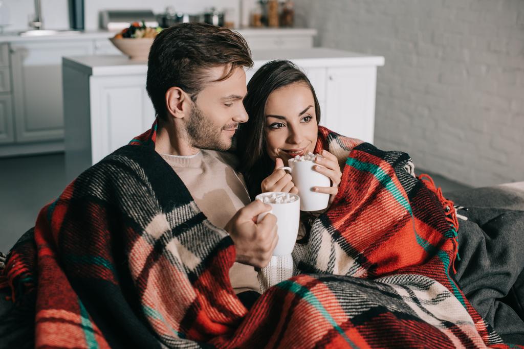 heureux jeune couple avec des tasses de cacao avec guimauve détente sur le canapé et couverture à carreaux
 - Photo, image