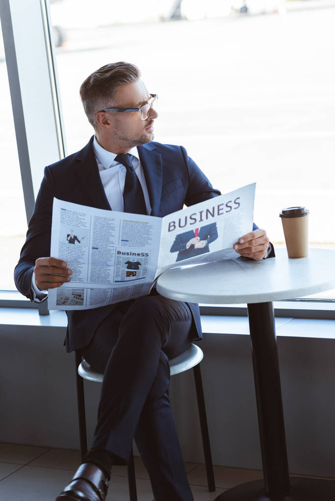 aikuinen liikemies lukee sanomalehteä ja katselee lentokentän ikkunaa
 - Valokuva, kuva