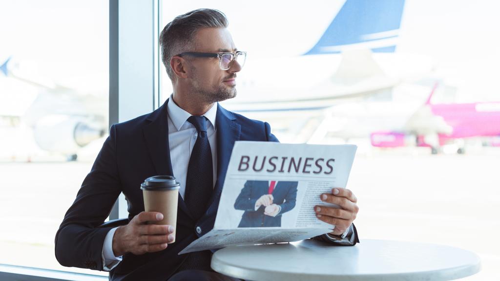adult handsome businessman with coffee to go reading newspaper and looking at window at airport - Photo, Image