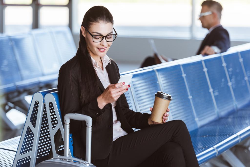 młody uśmiechający się interesu w okularach z kawą do za pomocą smartfona w odlotów Lotnisko - Zdjęcie, obraz