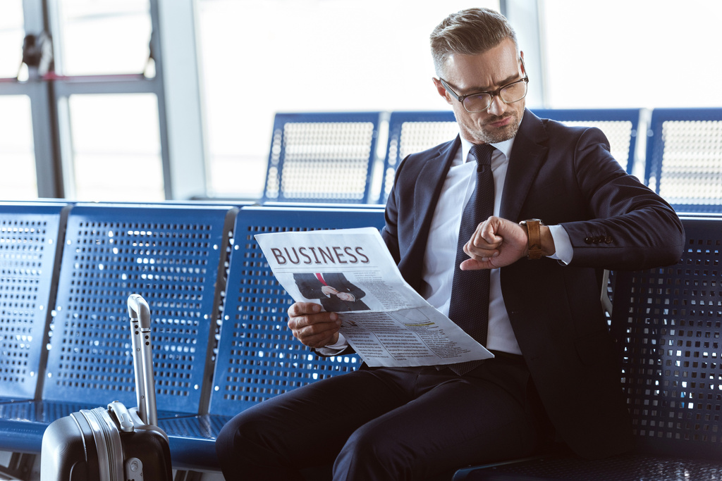 adult businessman sitting with newspaper at departure lounge at airport and looking at watch - Photo, Image