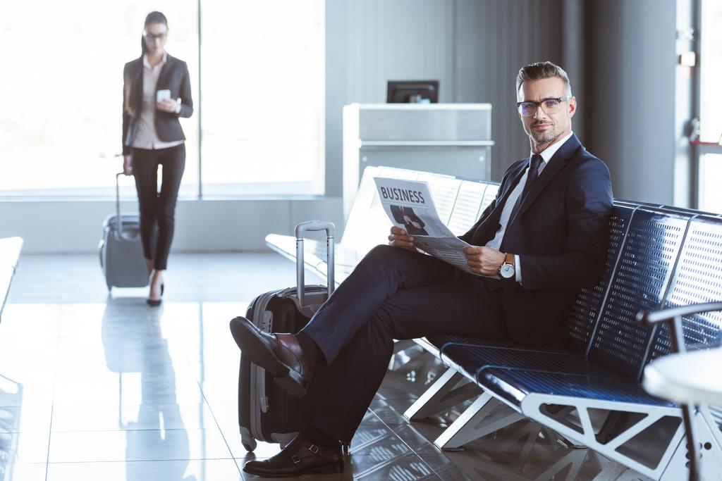 volwassen zakenman lezen van de krant terwijl zakenvrouw wandelen met bagage bij vertreklounge op luchthaven - Foto, afbeelding