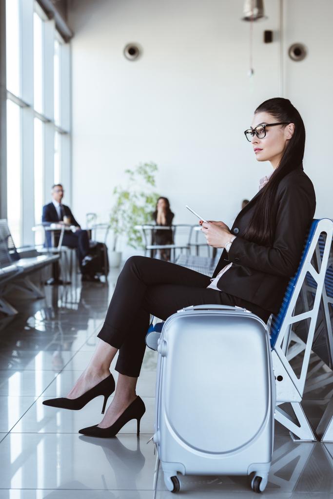 joven mujer de negocios en gafas con teléfono inteligente sentado en la sala de salida en el aeropuerto
 - Foto, imagen