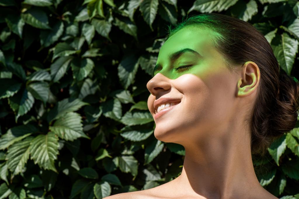 jonge lachende vrouw met groene schaduw op gezicht en laat bij achtergrond  - Foto, afbeelding