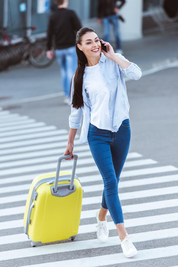 Kaunis hymyilevä nainen keltainen matka laukku ylitys jalankulkijoiden ja puhuu puhelimessa
 - Valokuva, kuva
