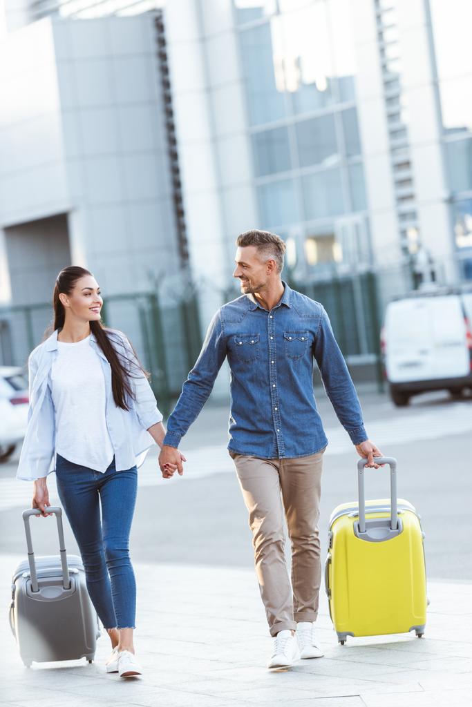 Пара туристов, держащихся за руки, вытаскивающих свой багаж, смотрящих друг на друга и улыбающихся на фоне аэропорта
 - Фото, изображение